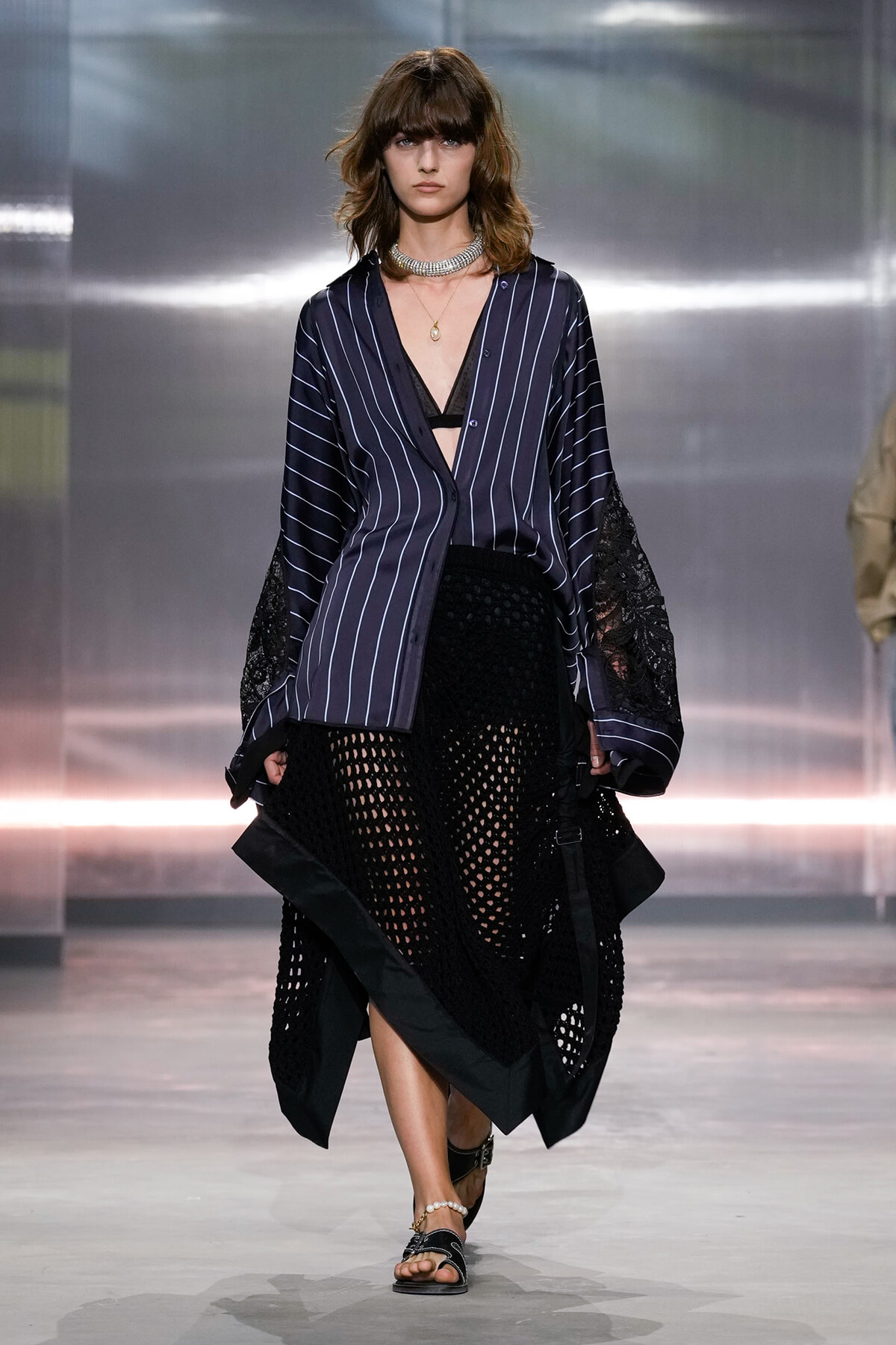 Model walking the runway in a navy pinstriped shirt with lace sleeves, a black crochet skirt, and flat sandals.
