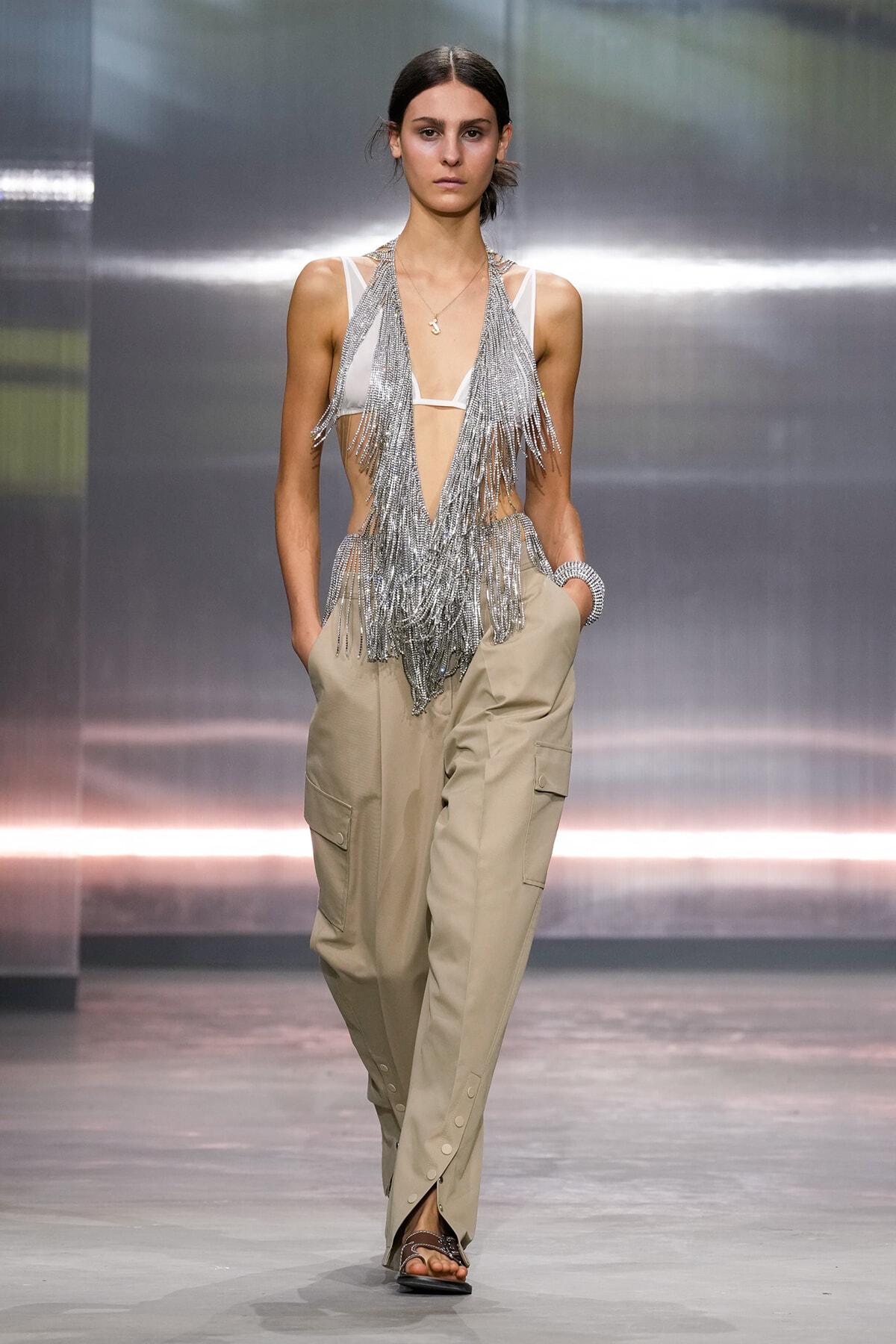 Model walking a runway in a white bralette with long silver fringe accessories and beige cargo pants, sandals visible beneath.