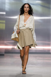 Model walking runway in a light striped blouse tucked into a tan A-line skirt, accessorized with layered necklaces and a tan perforated bag.