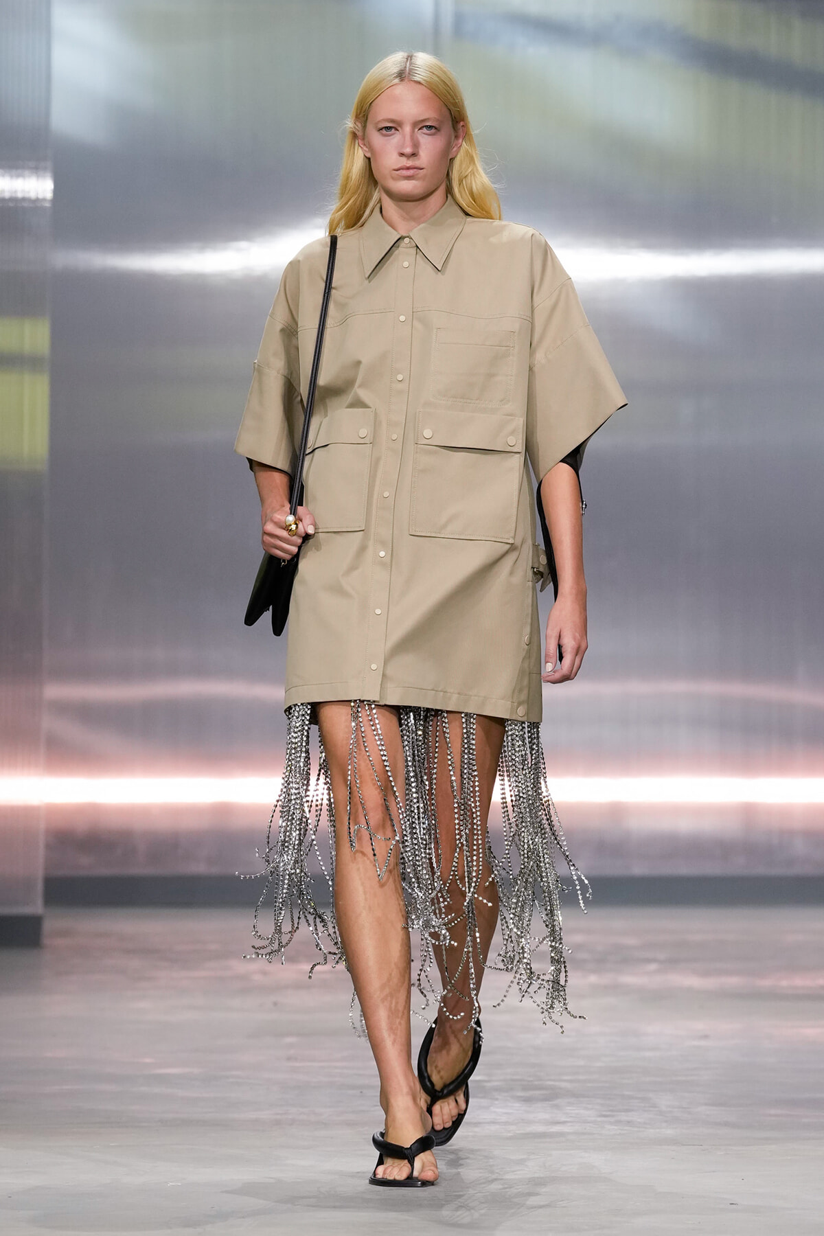 Model walking the runway in a beige oversized shirt dress with large front pockets and fringe chain details at the hem, paired with black sandals and a small black crossbody bag