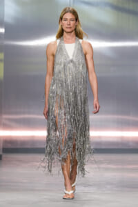 Model on a runway wearing a floor-length silver fringe dress and white sandals in a fashion show setting.