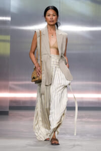 Runway model in a beige pinstripe sleeveless blazer with a tan bralette and white striped wide-leg pants, carrying a woven bag.