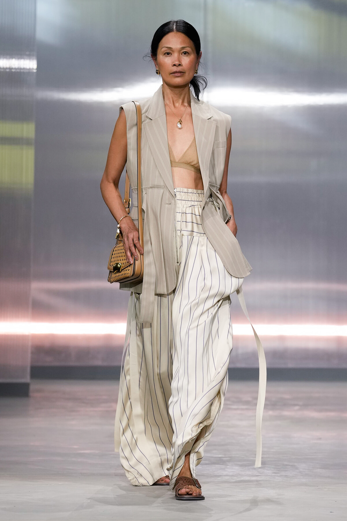 Runway model in a beige pinstripe sleeveless blazer with a tan bralette and white striped wide-leg pants, carrying a woven bag.