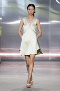 Model walking a runway in a pale green sleeveless V-neck dress with pockets, paired with white sandals and layered gold necklaces.