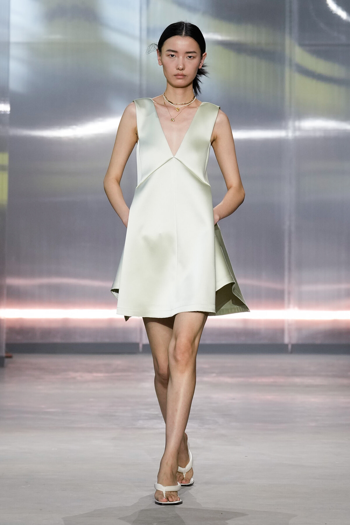Model walking a runway in a pale green sleeveless V-neck dress with pockets, paired with white sandals and layered gold necklaces.