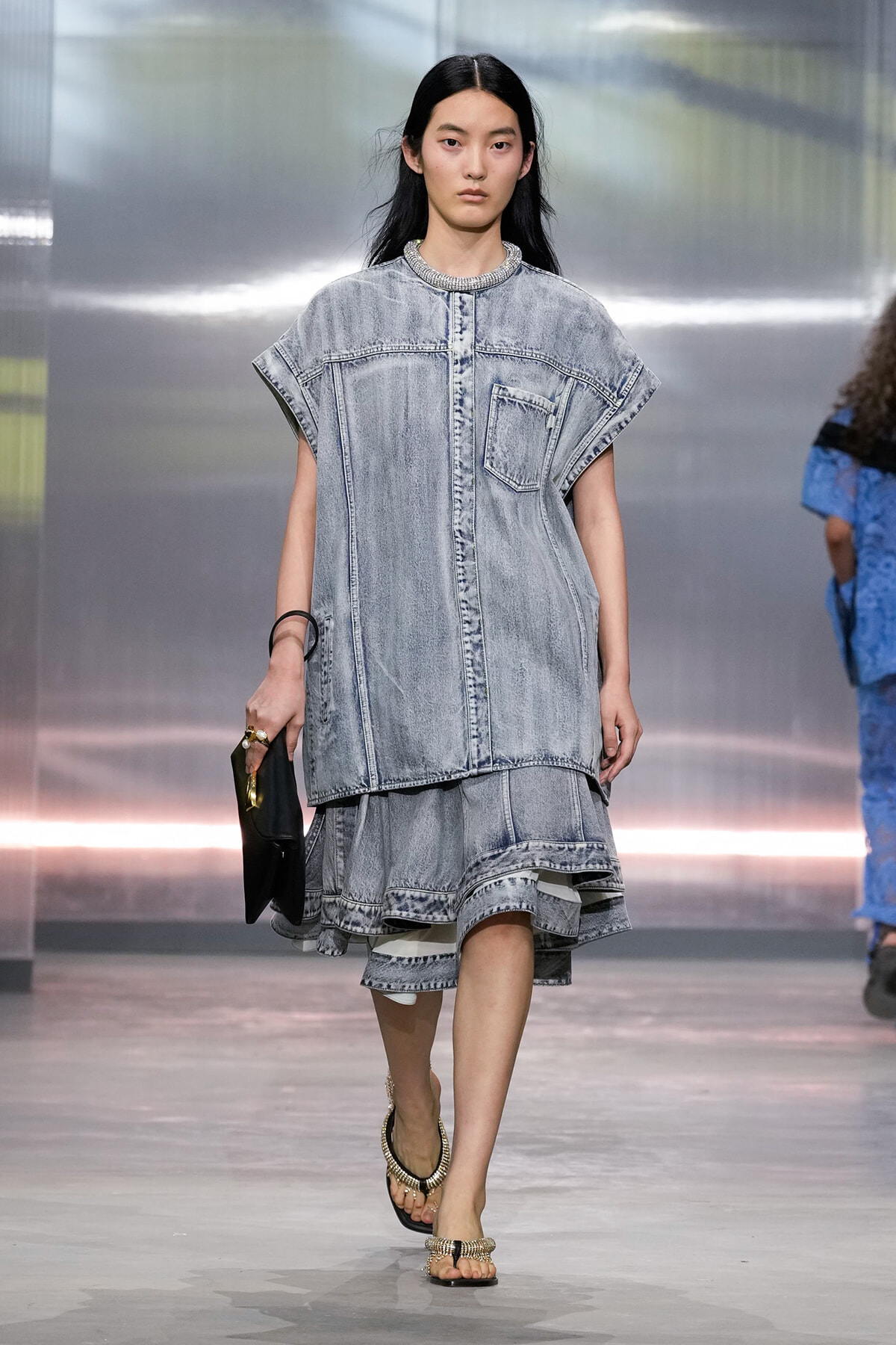 Model on a runway wearing an oversized light-wash denim tunic with a front pocket over a layered denim skirt, holding a black clutch and wearing sandals.