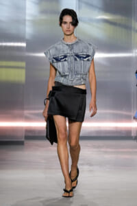 Model walks a runway in a cropped, light-wash denim top with large front pockets and a jeweled neckline, paired with a black asymmetrical wrap skirt and flat sandals.
