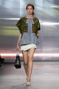 Model walking the runway in a camouflage top over a light denim vest, paired with a white mini skirt and white sandals, carrying a black mesh tote bag.