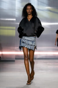 Model walking the runway in a black wrap-style top and a tiered, frilled denim mini skirt with sandals.