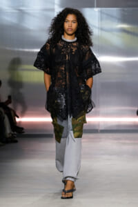 Female model walks the runway in a sheer black lace tunic over camo-panel gray cargo joggers and black sandals with a braided strap.