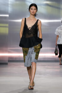 Model walks the runway in a black lace camisole over sheer top paired with patchwork denim shorts and gold sandals.