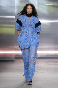 Runway model wearing a light blue lace ensemble with black bands on sleeves, walking toward camera with hands in pockets.