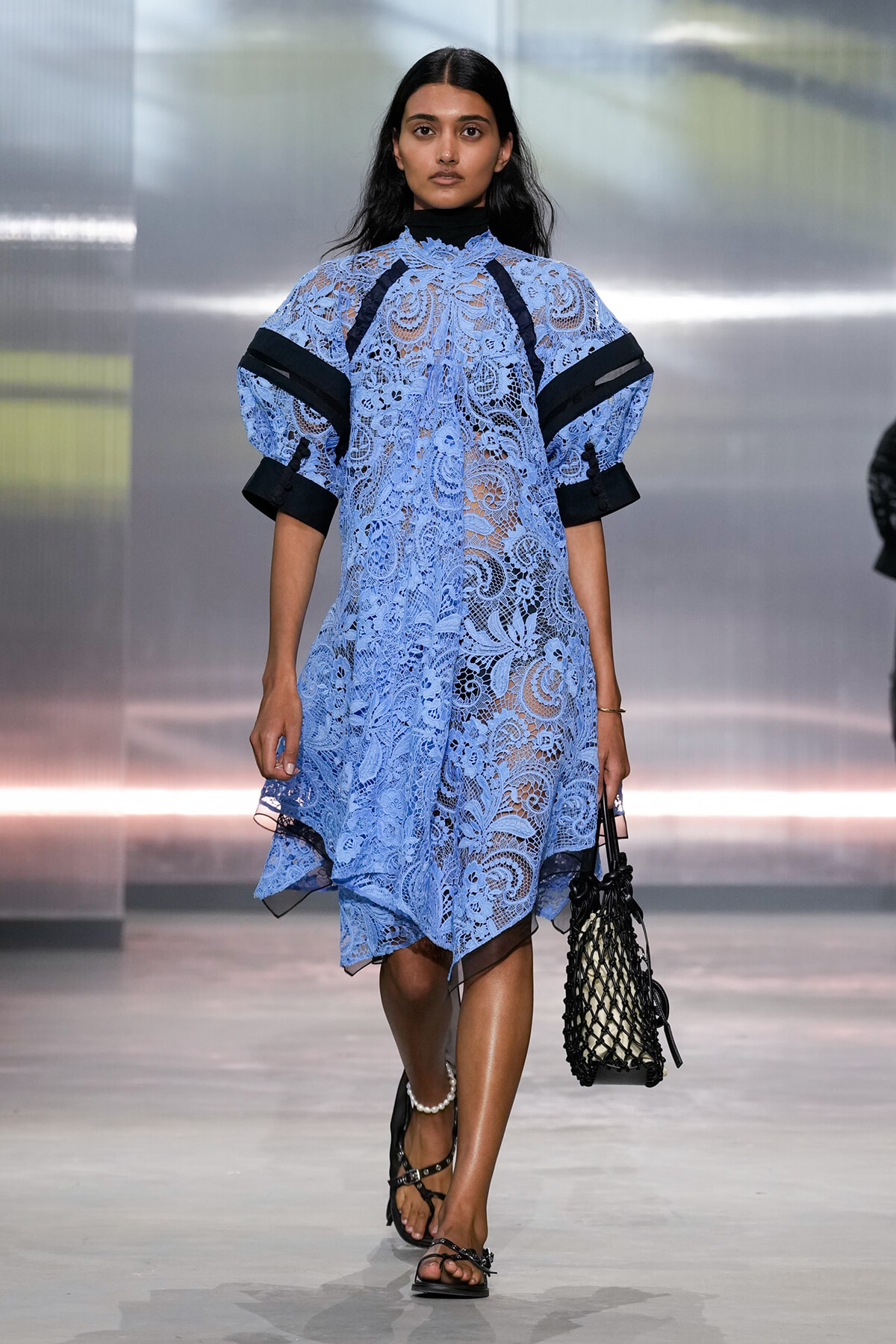 Model walks a fashion show wearing a light blue lace dress with voluminous sleeves and a black net handbag on her right arm, sandals on feet.