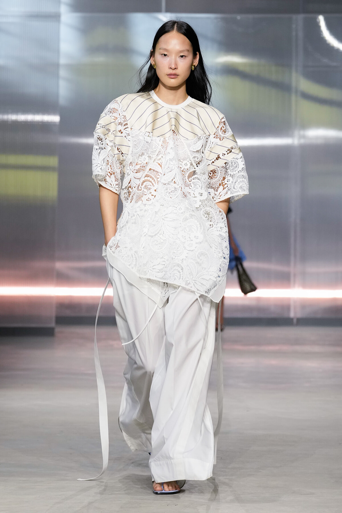 Model walking the runway in an all-white outfit: a loose lace blouse with a striped yoke paired with wide, flowing white pants.