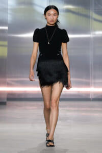 Model walking a runway in a black puff-sleeve top and feathered mini skirt, accessorized with a pendant necklace and flat sandals.