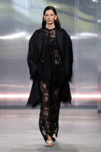 Model walks a runway in an all-black lace ensemble with a fringed coat and white sandals, gold earrings visible.