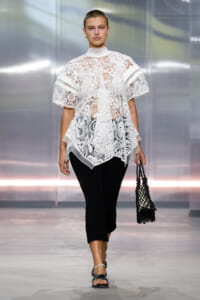 Model on a runway wearing a white lace top with a high neck and short sleeves over black cropped pants, holding a black woven handbag.