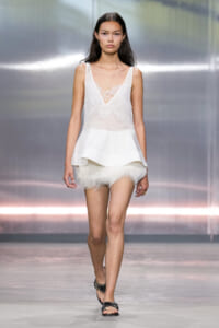 Model walking a runway in a white lace-trim sleeveless top with a layered peplum and white feathered shorts, wearing black sandals.