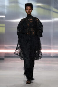Model walks the runway in a black lace oversized top with voluminous sleeves and feathered black pants on a fashion show set.