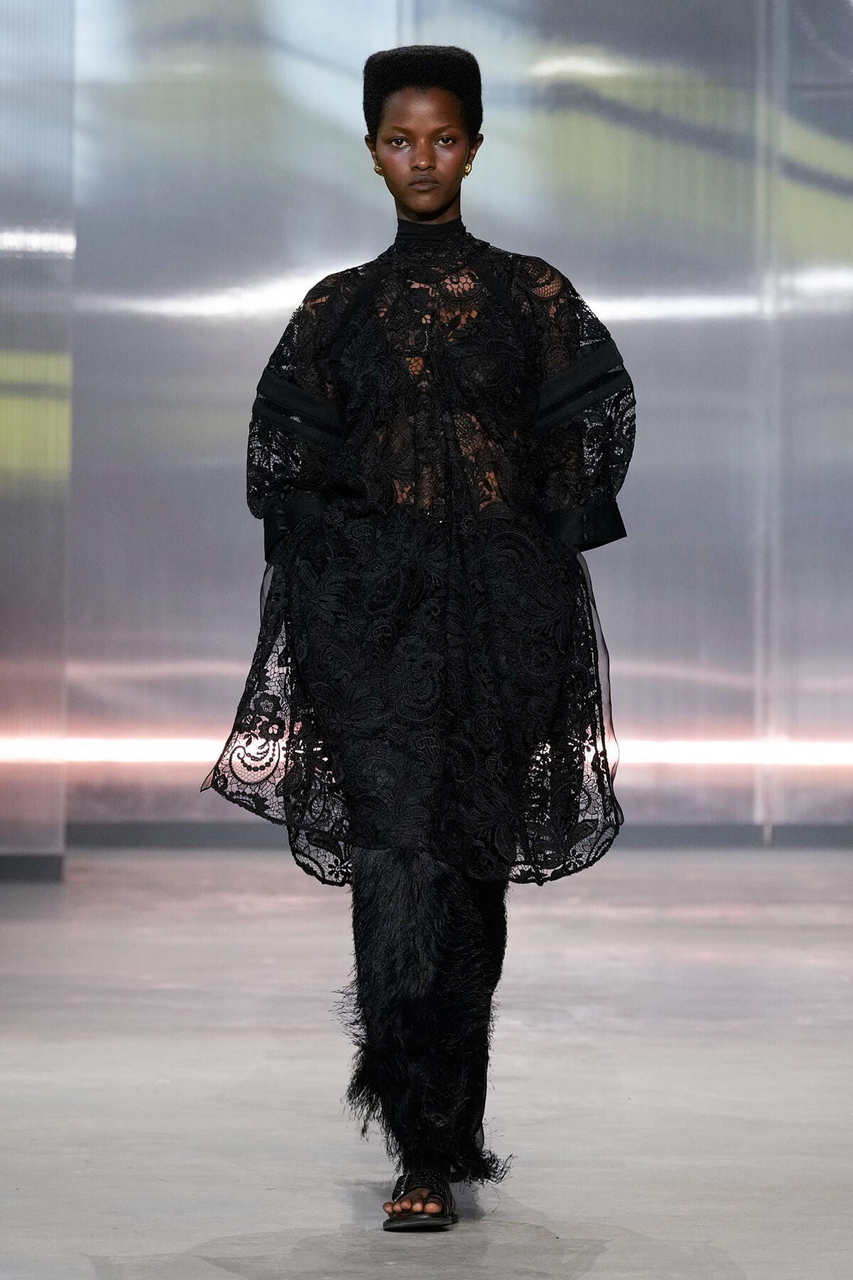 Model walks the runway in a black lace oversized top with voluminous sleeves and feathered black pants on a fashion show set.
