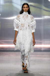 Model walks the runway in a white lace high-neck dress with puff sleeves and scalloped hem.