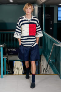 Male model walking down a stairwell in a subway station, wearing a navy-and-white striped knit sweater with a red square panel, navy shorts, and dark blue socks.