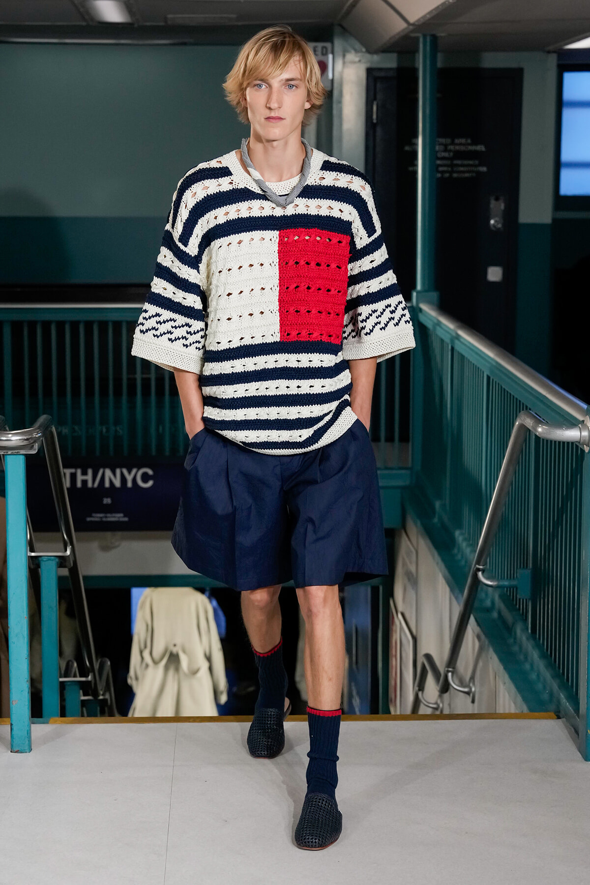 Male model walking down a stairwell in a subway station, wearing a navy-and-white striped knit sweater with a red square panel, navy shorts, and dark blue socks.