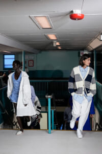 Two models in blue-and-white outfits walk down a staircase runway in a transit station setting.