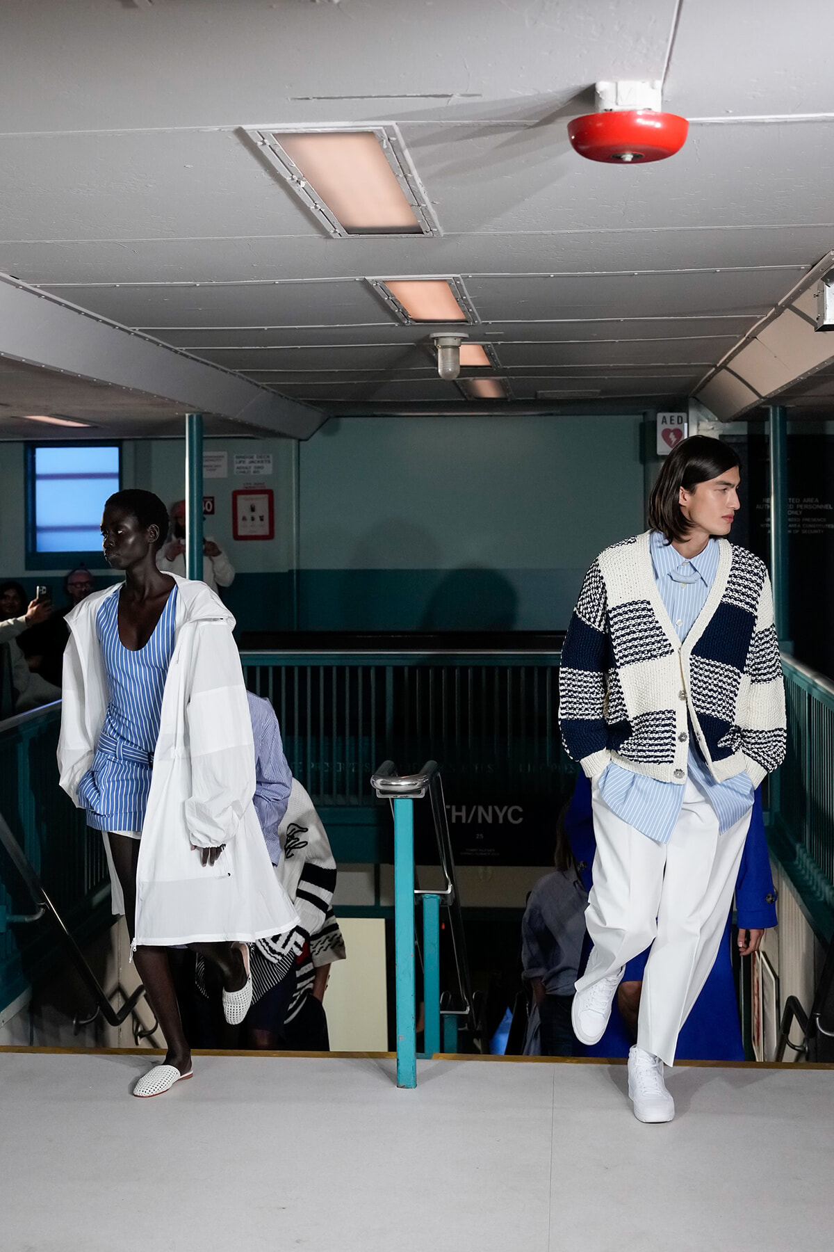 Two models in blue-and-white outfits walk down a staircase runway in a transit station setting.