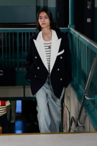 Young person walking in a subway station wearing a navy double-breasted blazer with white lapels, striped tee, and light blue jeans.