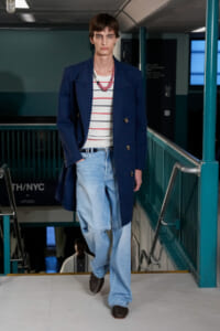 Male model walking down a stairwell runway in a navy double-breasted coat, striped tee, and light-wash jeans.