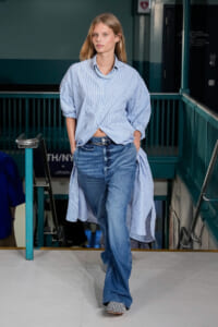 Model walks the runway in an oversized blue striped shirt tied at the waist and wide-leg jeans, hands in pockets.