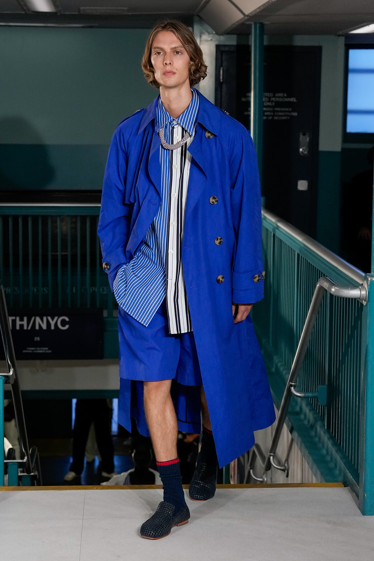 Male model in a bright blue trench coat over a blue-and-white striped shirt, walking on a subway platform runway with teal railings and a chunky silver necklace