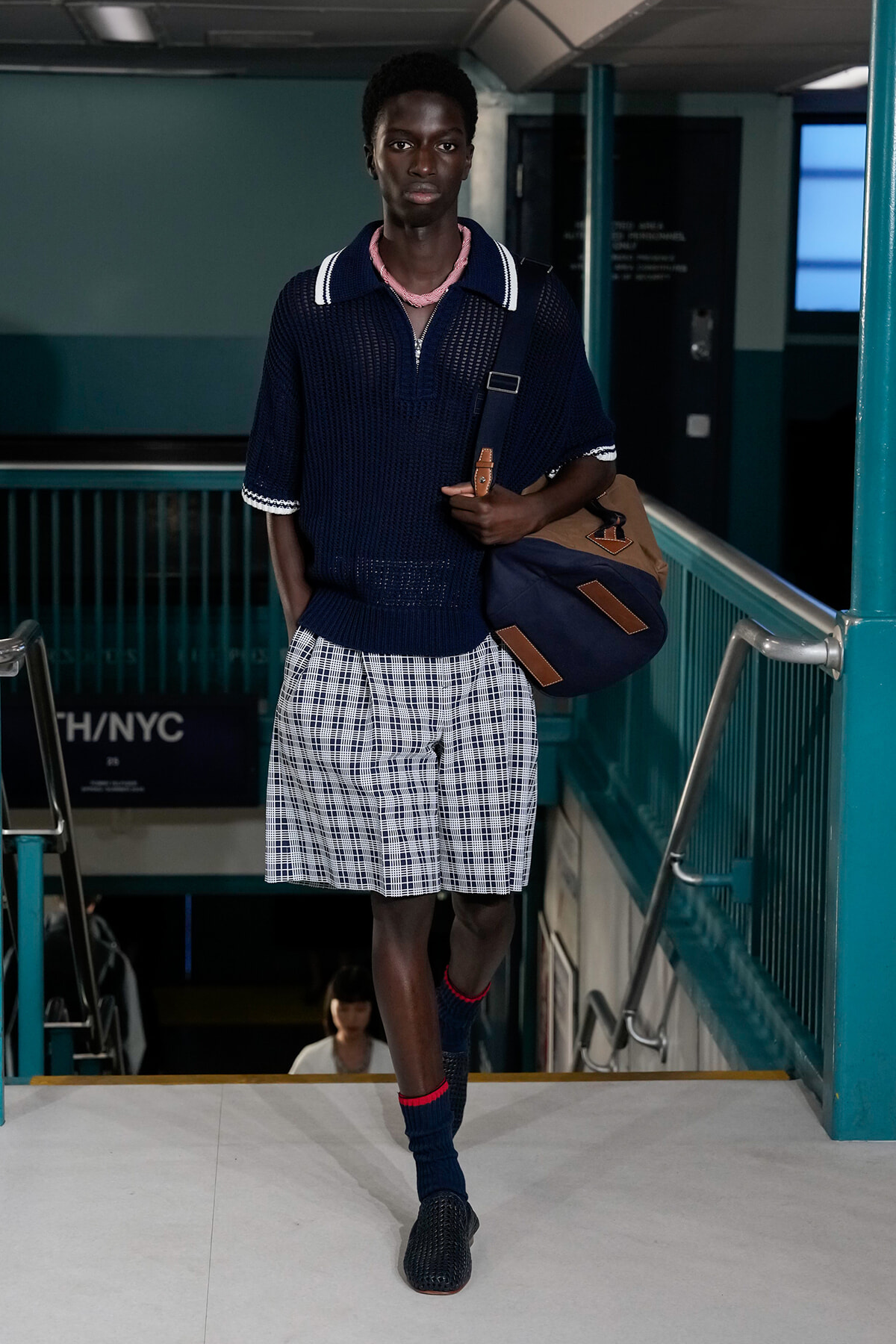 Man wearing a navy knit polo and plaid shorts carries a brown-and-navy duffel bag while walking down subway stairs in a station.