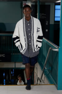 Male model descending a stairwell in a navy-striped shirt and oversized white cardigan with navy trim, navy shorts, during a fashion show.