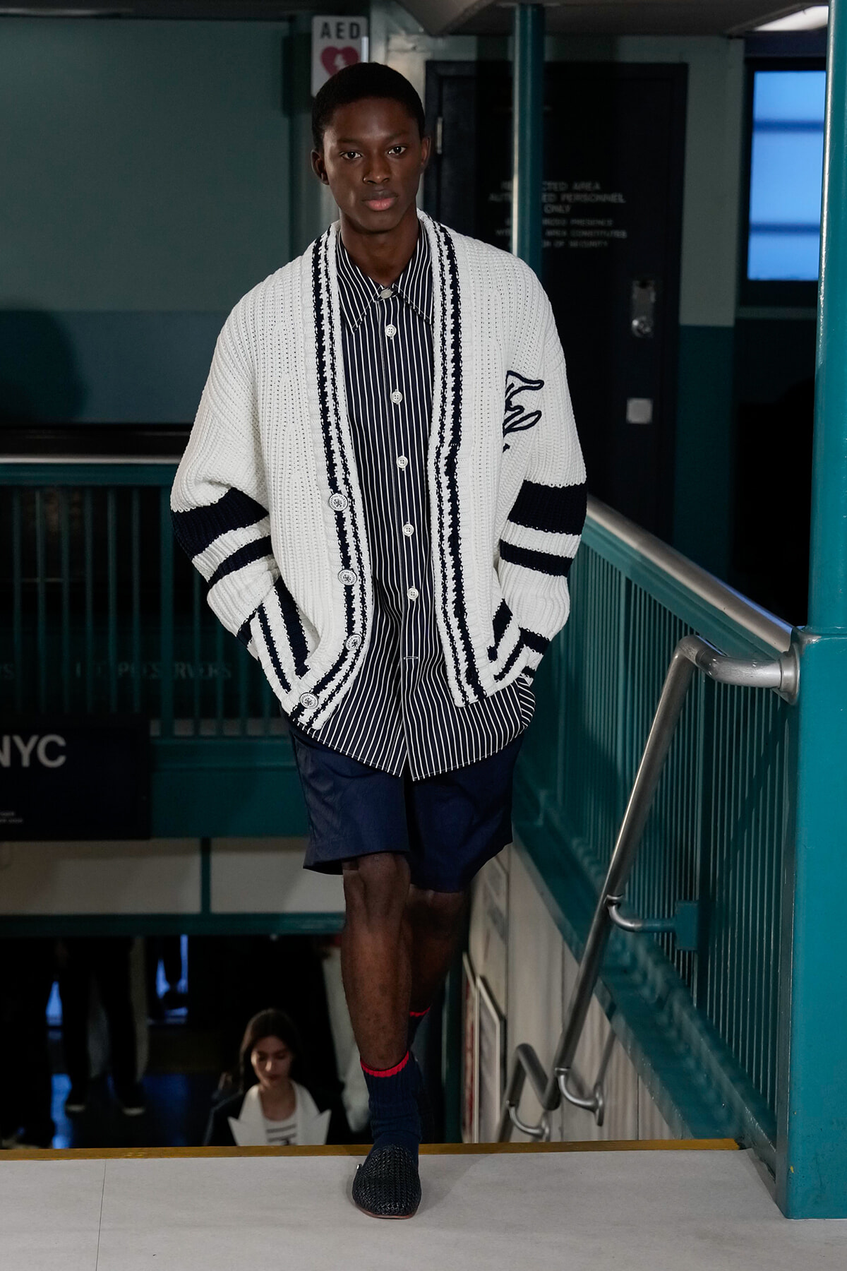 Male model descending a stairwell in a navy-striped shirt and oversized white cardigan with navy trim, navy shorts, during a fashion show.