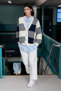Male model walking down a subway stairwell in a chunky navy-and-white striped cardigan over a light blue striped shirt and white trousers, with white sneakers.