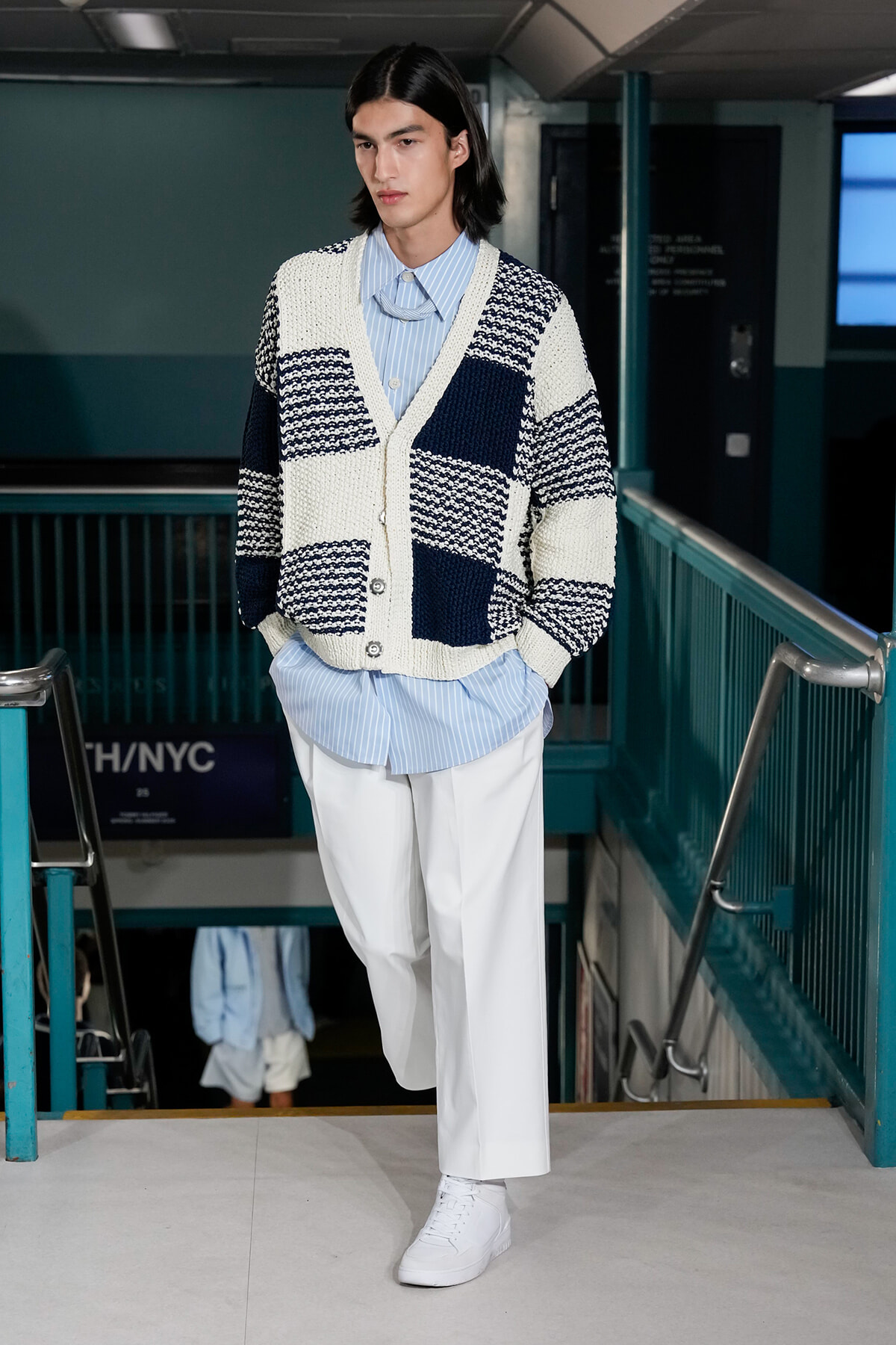 Male model walking down a subway stairwell in a chunky navy-and-white striped cardigan over a light blue striped shirt and white trousers, with white sneakers.