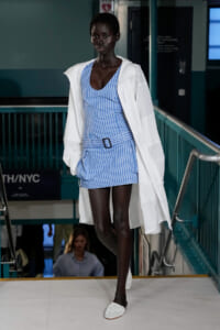 Model wearing a blue striped belted mini-dress with a white oversized jacket, walking a runway.