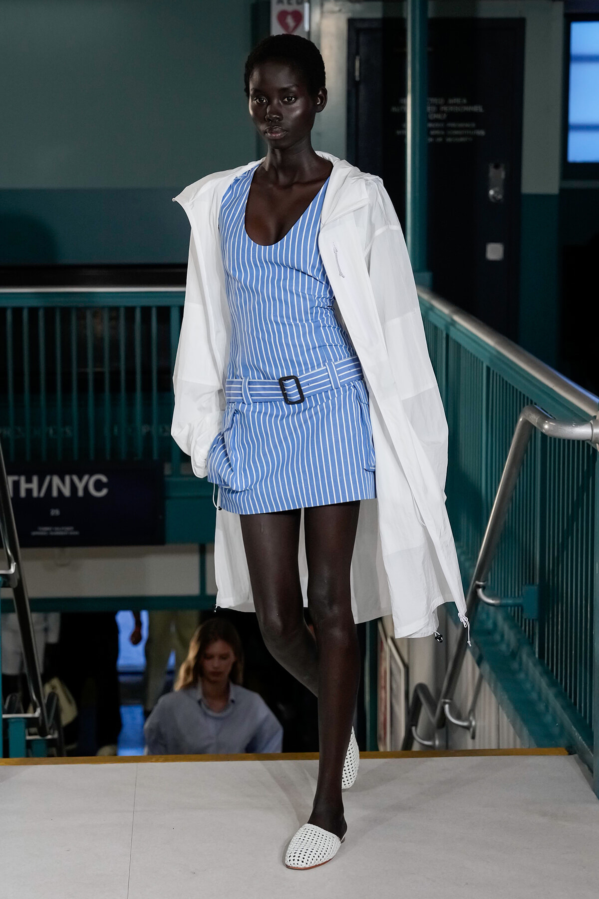 Model wearing a blue striped belted mini-dress with a white oversized jacket, walking a runway.