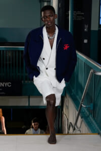 Male model walks down a runway wearing a navy jacket over a white double‑breasted suit and a chunky silver necklace.