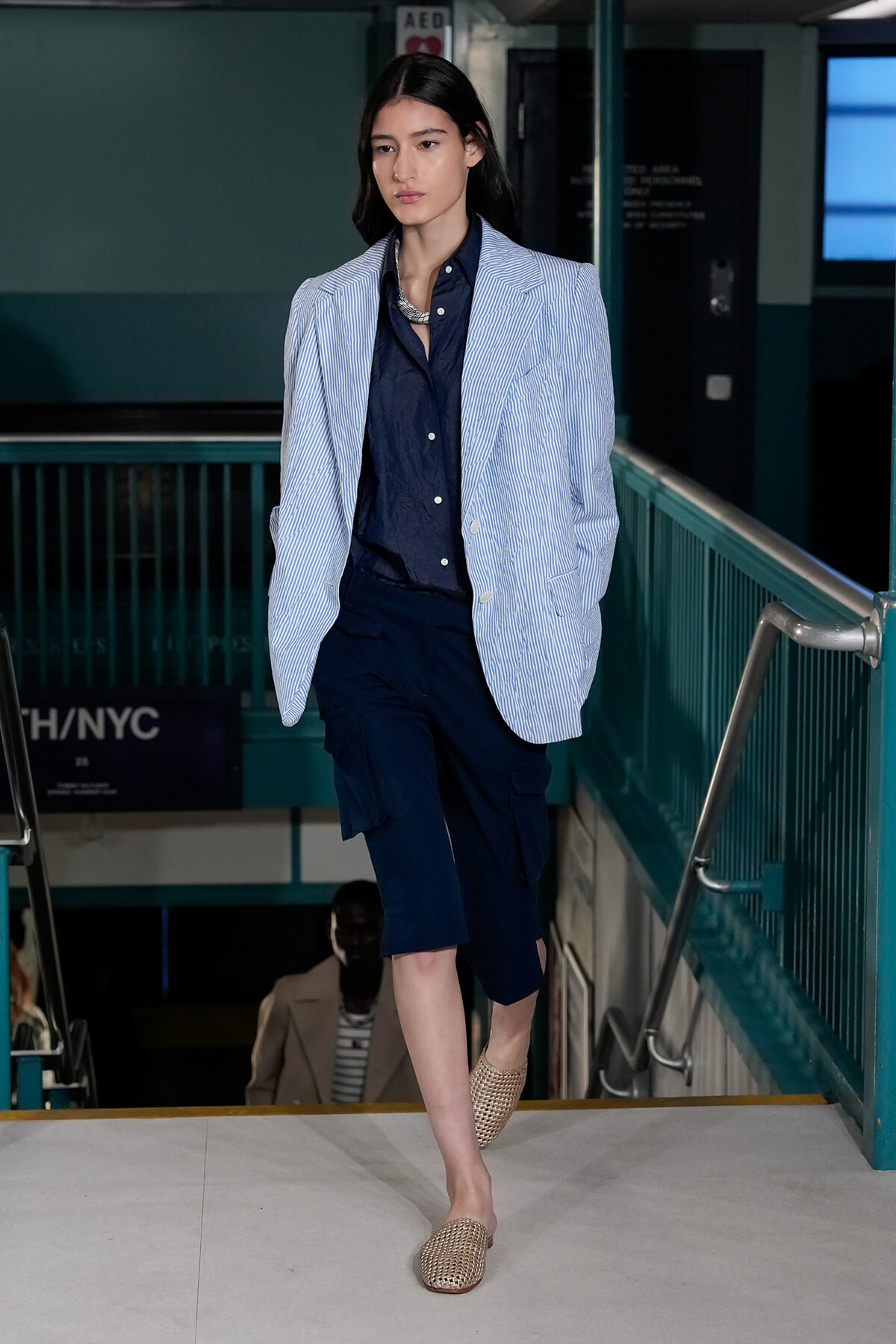 Model walking down a runway in a navy shirt with a light blue striped blazer and navy cargo shorts, paired with woven tan mules.