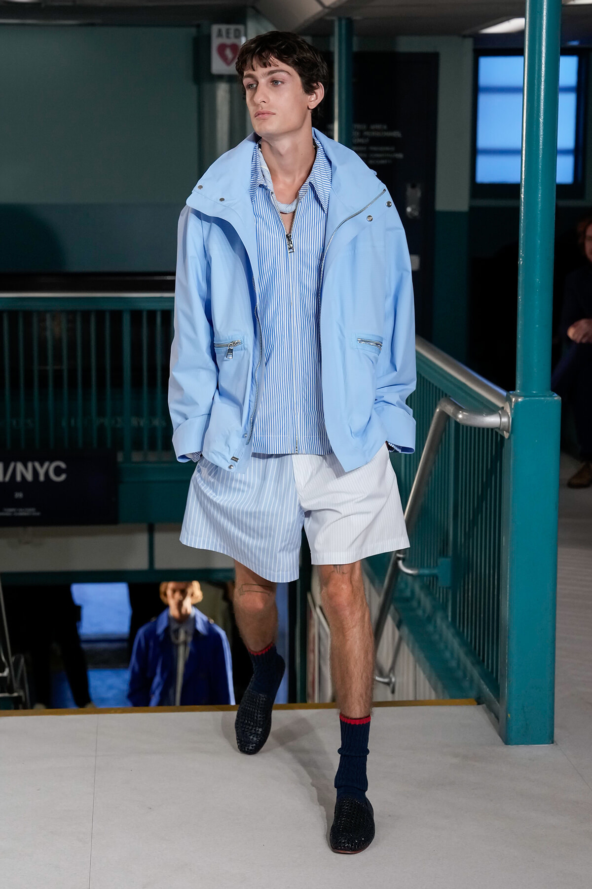 Male model walking down a runway in a light blue jacket, blue striped shirt, and light shorts, with dark socks and dark shoes in a subway-station set-up