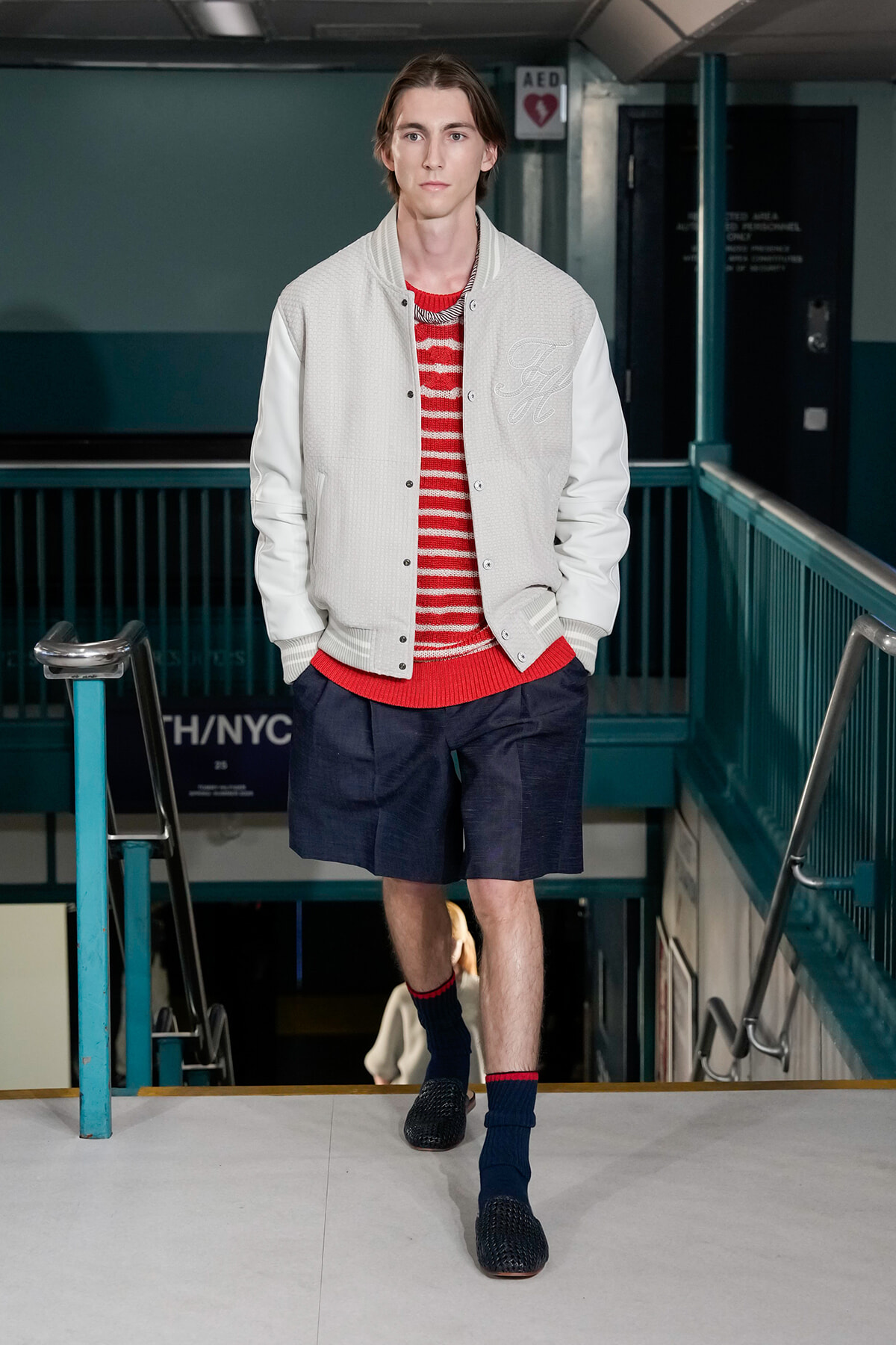Male model walking down a teal subway stairwell in a light varsity jacket over a red striped sweater, navy shorts and blue socks.