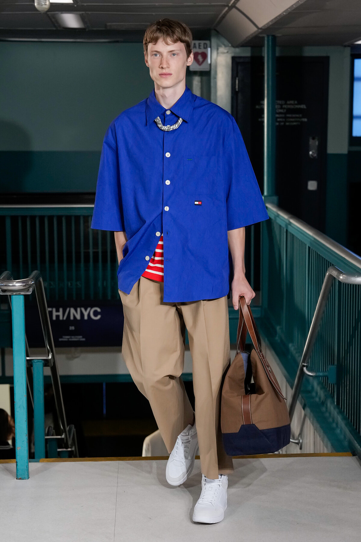 Male model wearing an oversized royal blue shirt, beige trousers, and white sneakers, carrying a brown duffel bag on a runway.