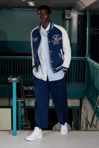 Model in navy varsity jacket with white sleeves, light blue striped shirt, wide-leg navy pants, and white sneakers standing on a subway platform stairwell.