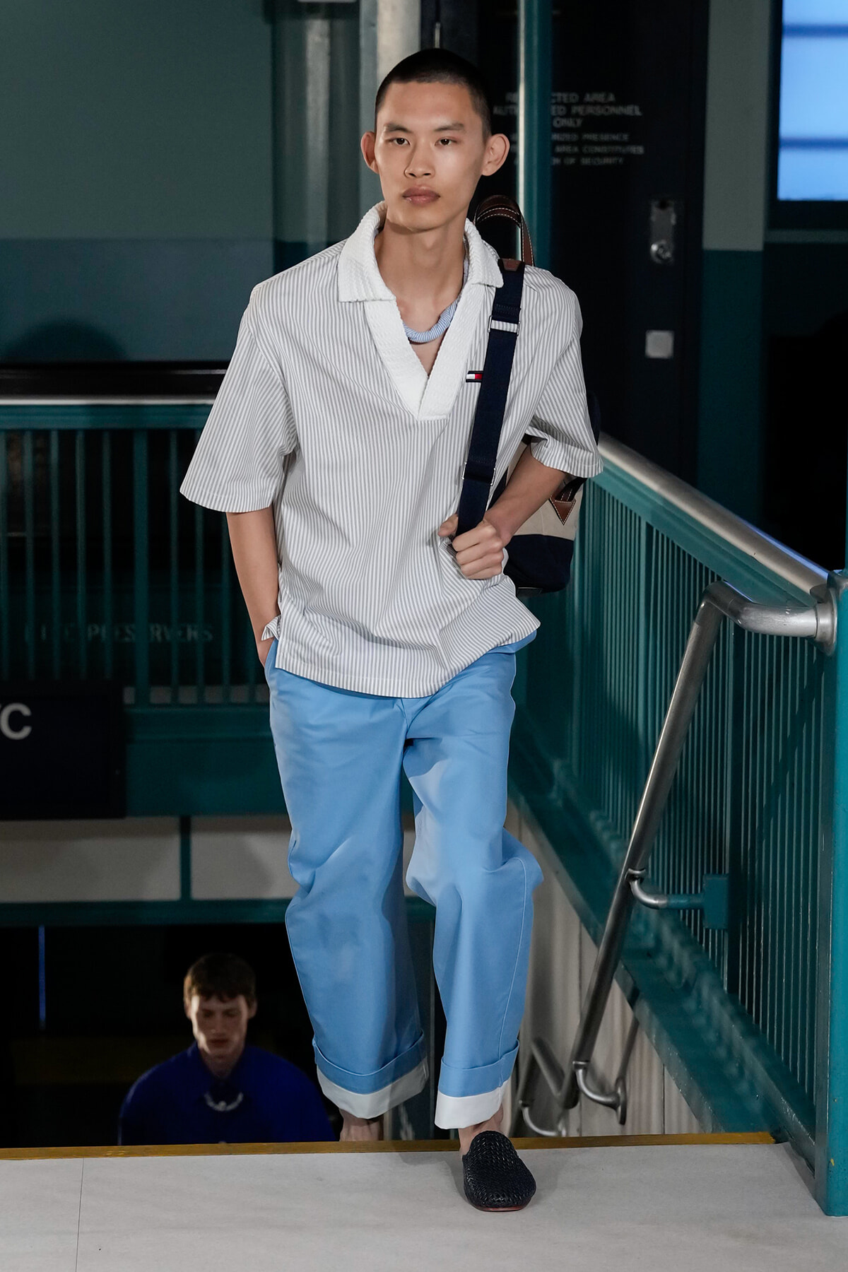 Young man wearing a striped white shirt and light blue pants walking down stairs with a backpack over one shoulder in a tunnel-like hallway.