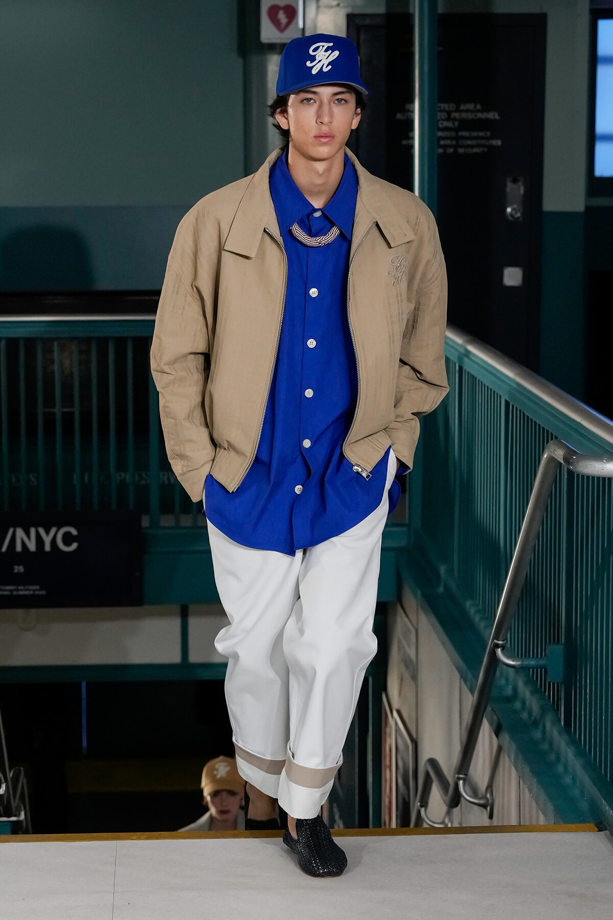 Male model in a blue shirt and cap wearing a beige jacket and white cropped pants walks down a stairwell.