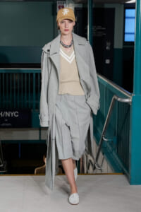Model on a runway wearing a light gray oversized suit with matching shorts, beige V-neck sweater, and tan cap in a subway-station setting.] ,