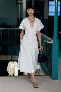 Model in a white V-neck midi dress with short sleeves walks down a runway stair, carrying a navy handbag and wearing a chunky necklace.